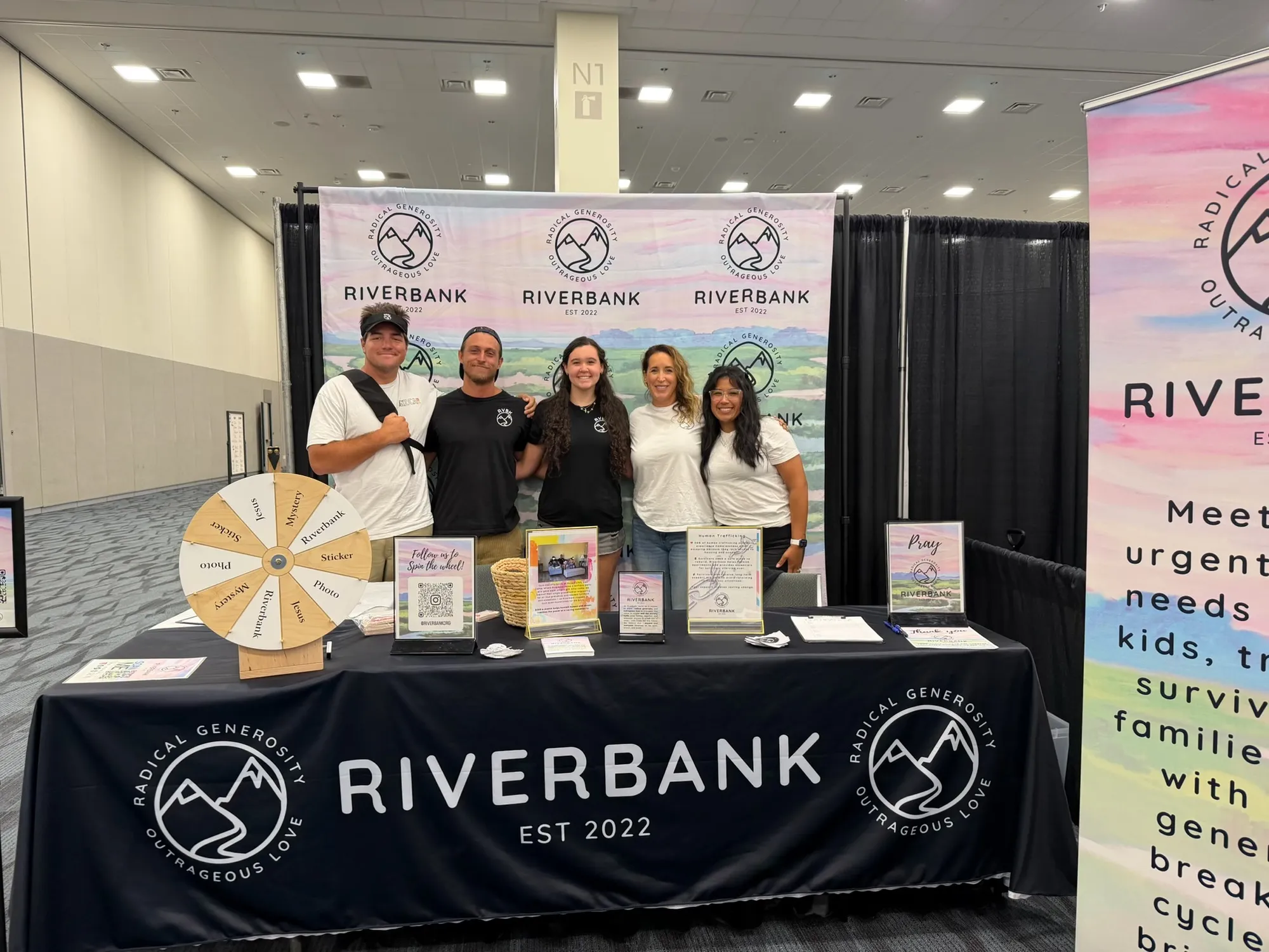 Five people behind a Riverbank booth table at a convention hall, with the Riverbank logo on the tablecloth, a prize wheel, and a printed backdrop behind them