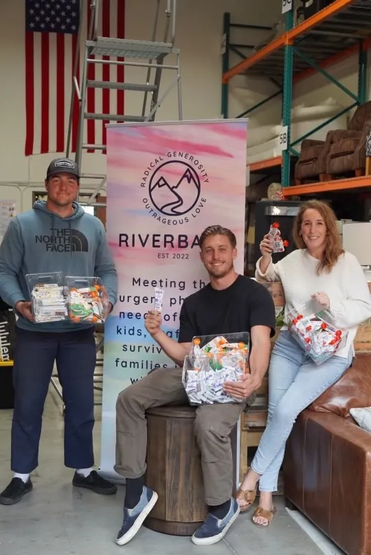 Three people holding clear bins of packed supply kits in front of a Riverbank banner reading radical generosity, outrageous love