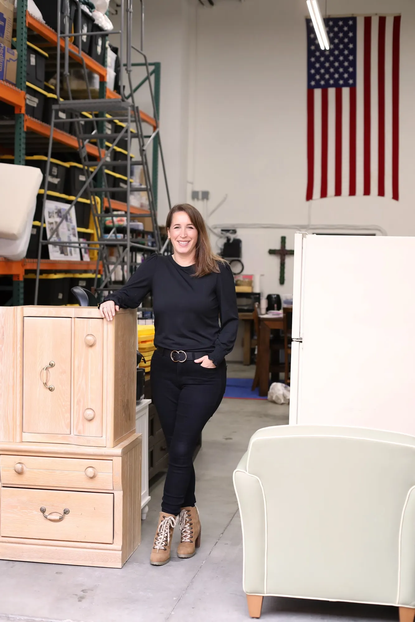 Nadia Caporaso standing beside a stacked wooden dresser in the Riverbank warehouse, with pallet racking, a refrigerator, and an armchair around her