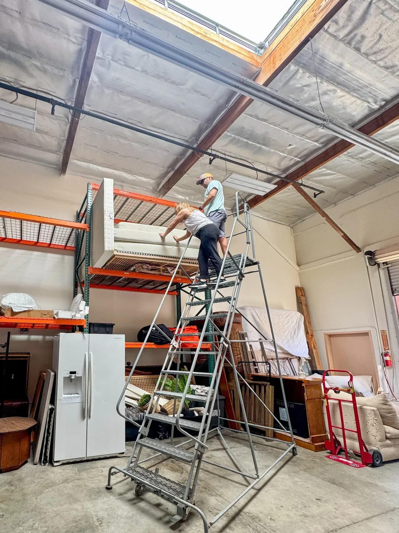Two people handling a mattress on a rolling ladder beside the pallet racking in the Riverbank warehouse