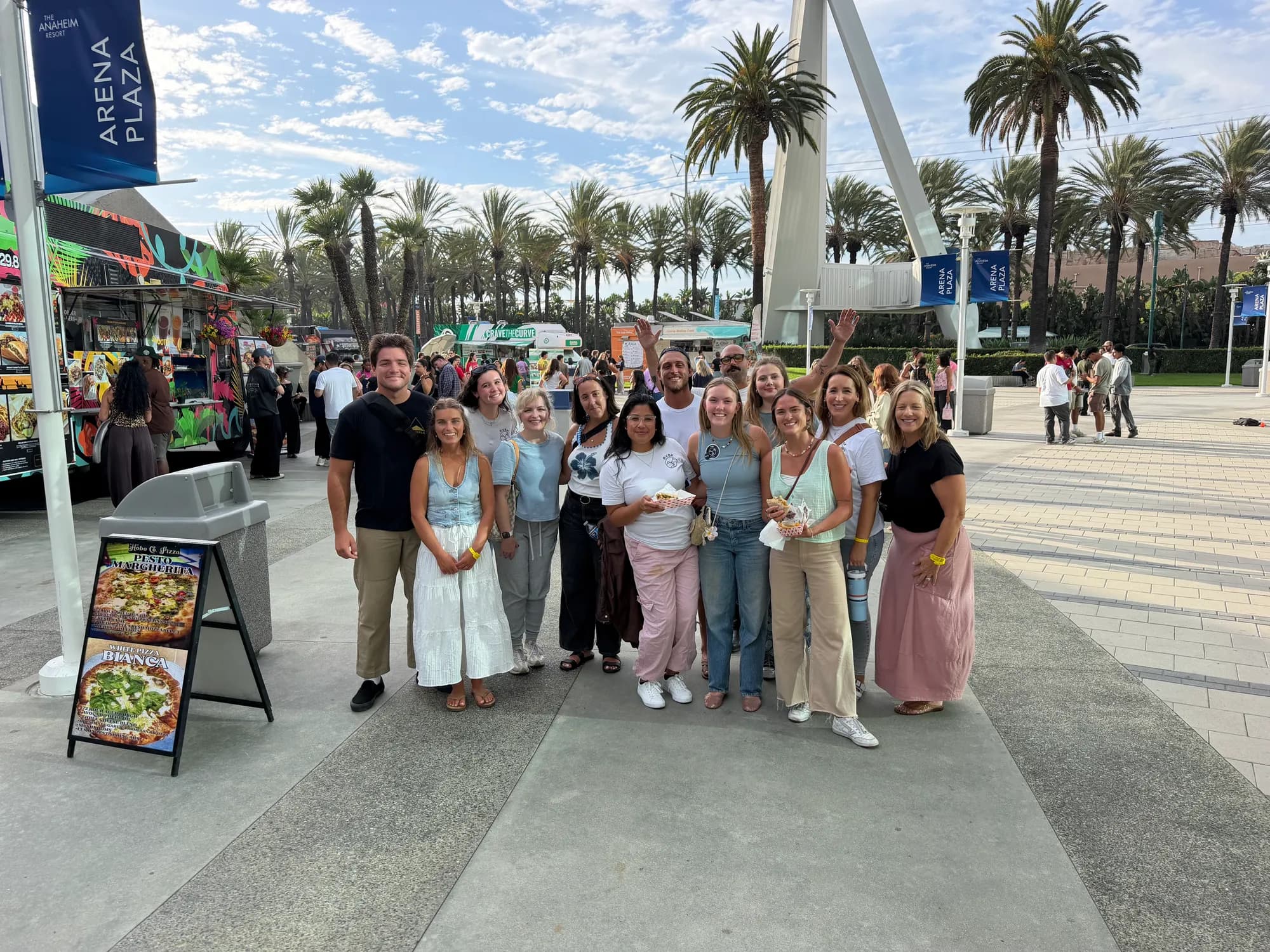 A large group standing together on a sunny plaza lined with palm trees and food trucks, two of them waving