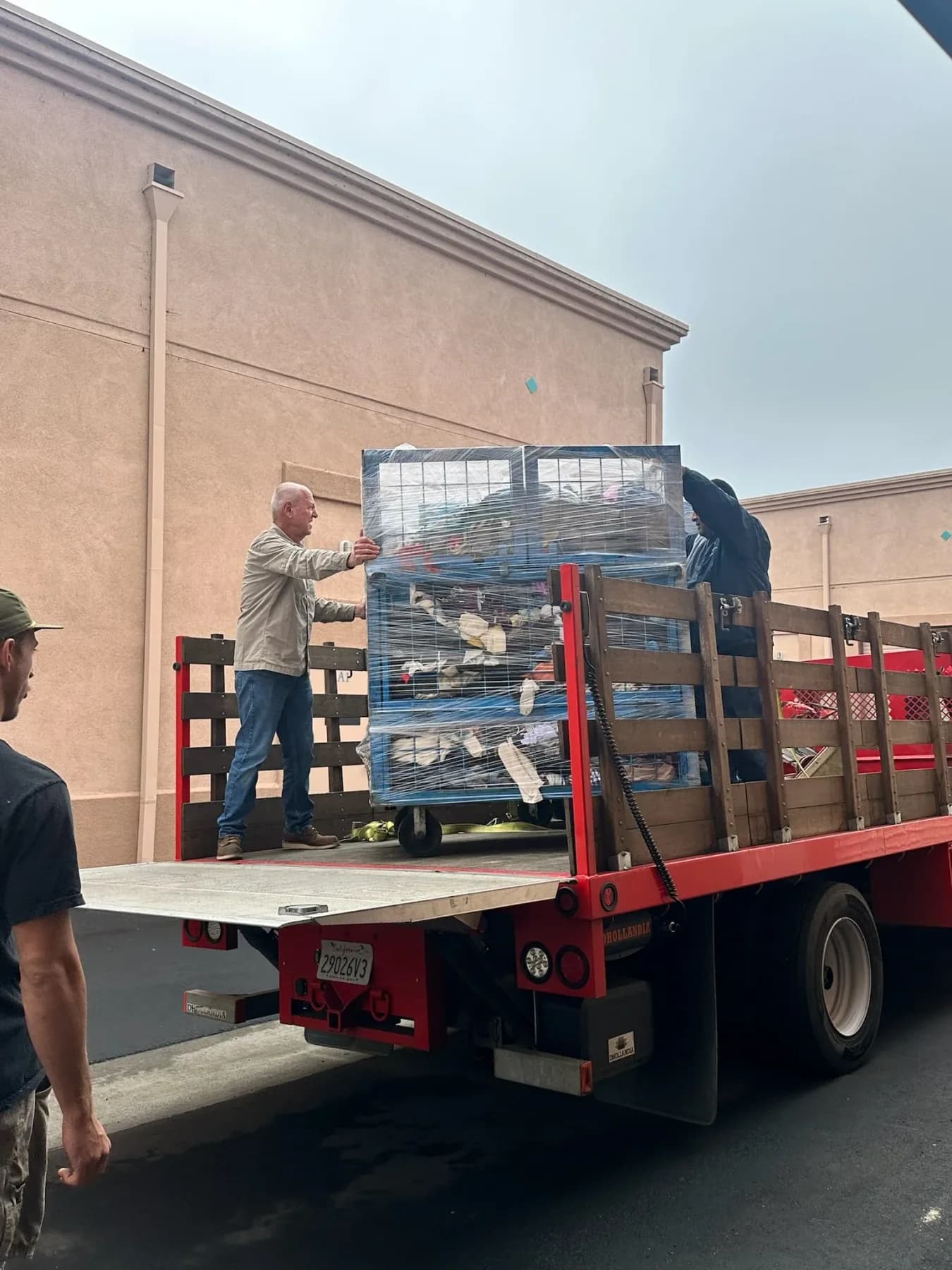 Two people steadying a shrink-wrapped cage cart on the deck of a red stake-bed truck outside a commercial building