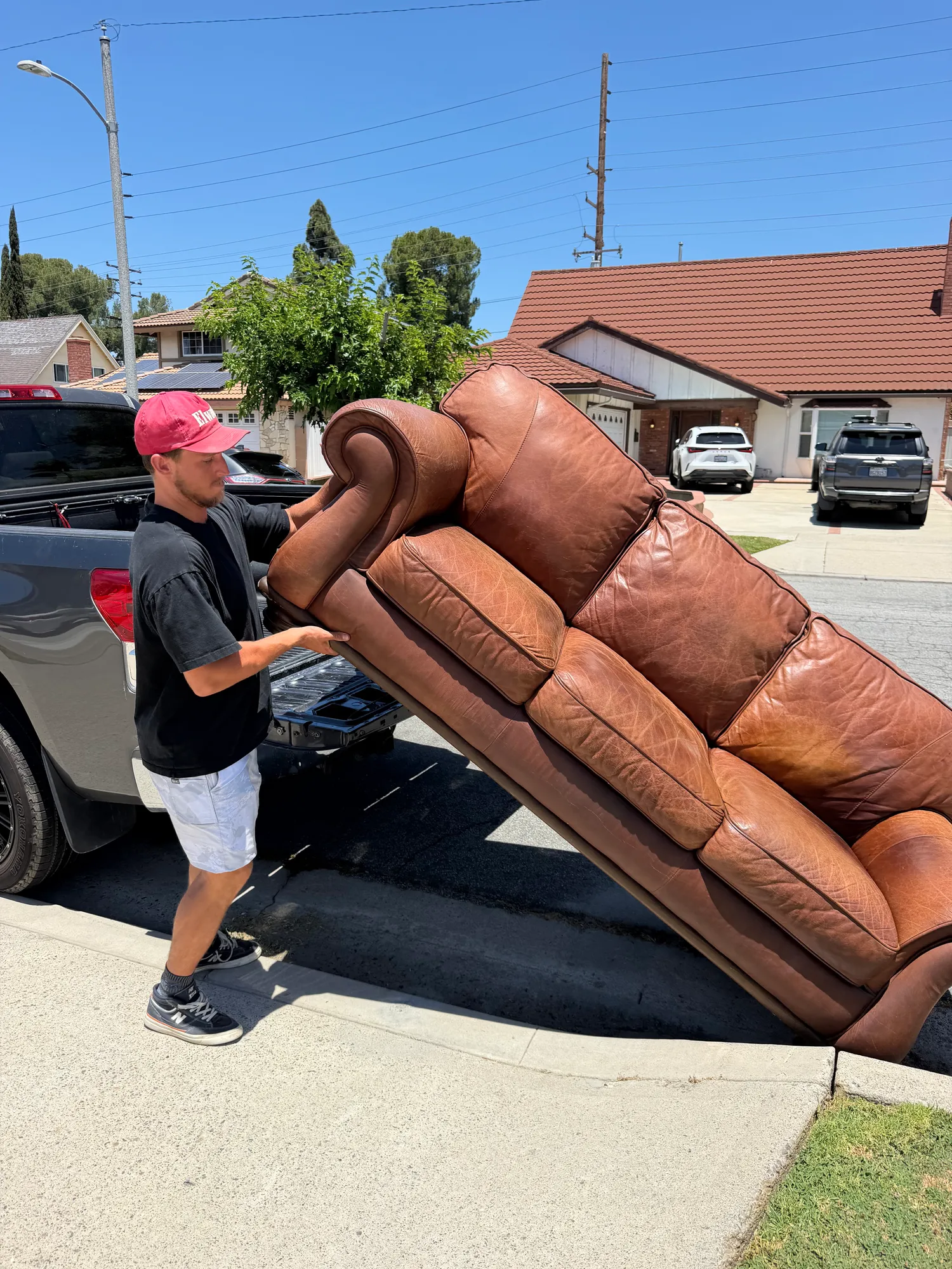 A man tipping a brown leather sofa into the bed of a pickup truck at the curb outside a house