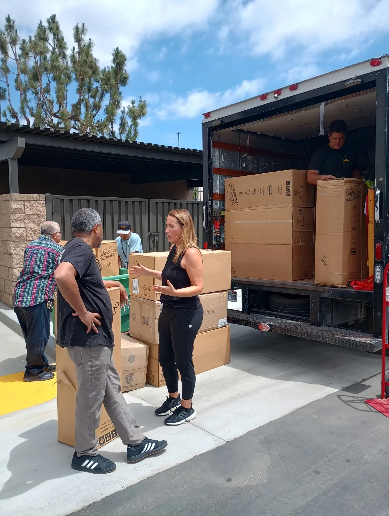 A box truck backed up outside a building with mattress cartons stacked inside, and a woman in black talking with two men beside more boxes on the pavement
