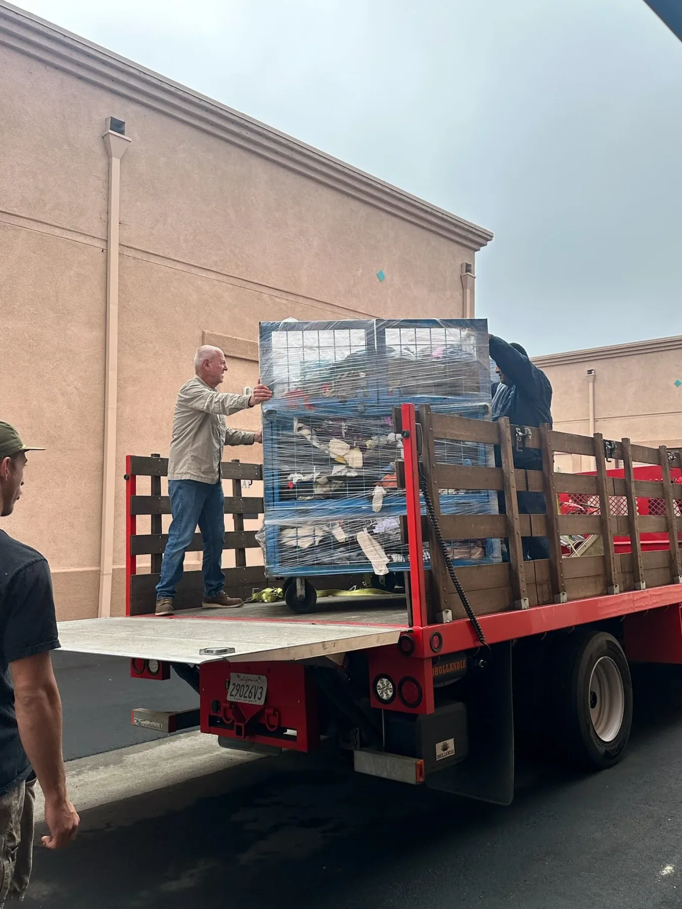 Two people steadying a shrink-wrapped cage cart on the deck of a red stake-bed truck outside a commercial building